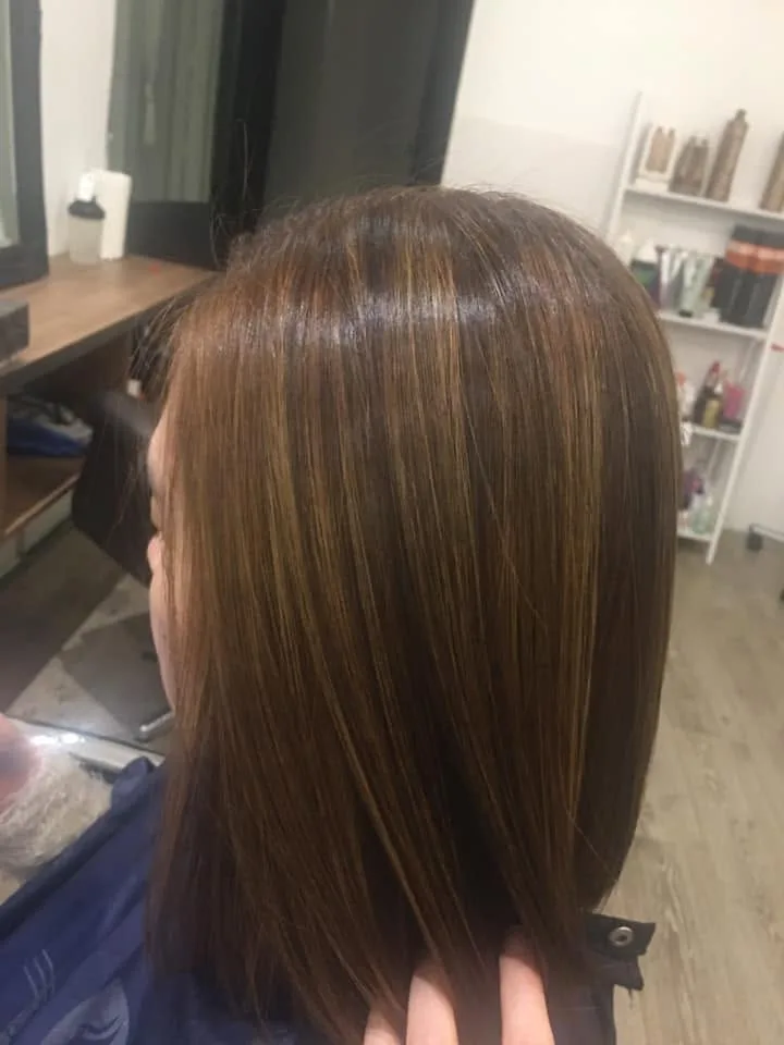 Glossy caramel-highlighted bob in profile — white retail shelf of product bottles, timber bench and mirror