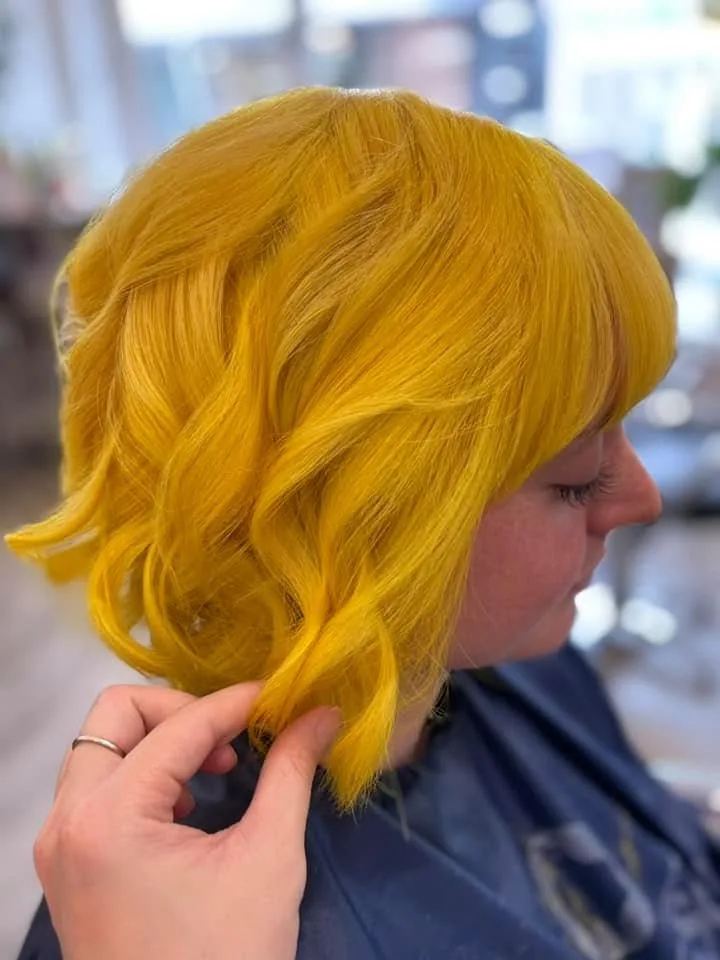 Yellow bob profile — stylist's hand lifting a curl at the nape, navy cape