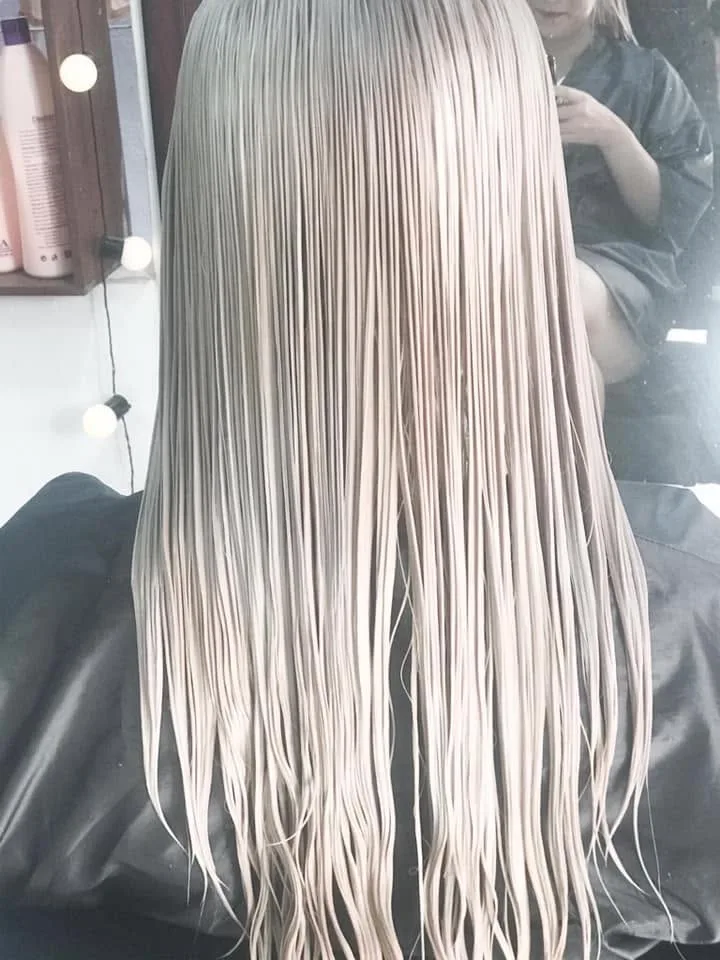 Very long platinum-blonde hair combed straight — bulb-lit vanity, client in black cape