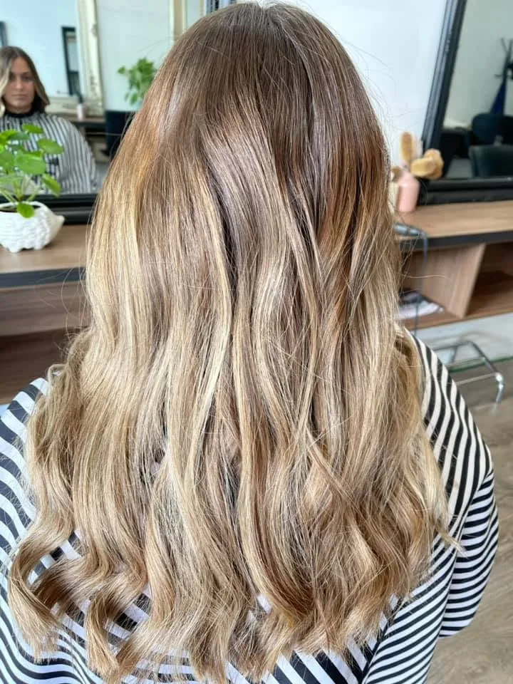 Long honey balayage waves — timber bench, monstera in white pot, large gilt mirror reflecting the salon